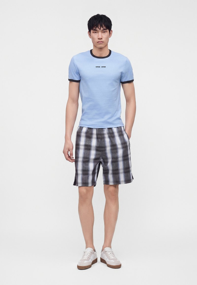 Homme portant un t-shirt bleu clair avec bordure noire et texte, un short à carreaux noir et blanc, et des baskets blanches, debout devant un fond uni.