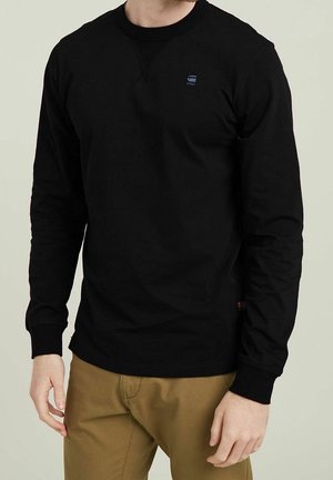 Homme portant un shirt à manches longues noir avec un petit logo sur la poitrine gauche et un pantalon kaki, debout devant un fond neutre.
