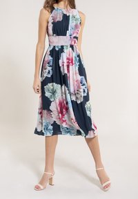 Robe midi florale avec un fond marine, des fleurs pastels roses et bleues, un corsage plissé et une taille assortie. Associée à des sandales à talons blocs.