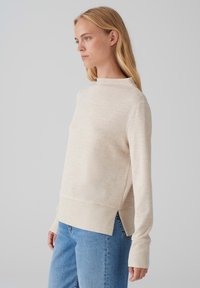 Light beige sweater with a high neckline and ribbed hem. Features a loose fit, long sleeves, and side slits. Made from soft knit material.