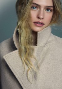 Bež volnen plašč z visokim ovratnikom, s čistim, kotnim dizajnom. Dolgi, valoviti blond lasje delno obkrožajo obraz. Mehak teksturni detajl.