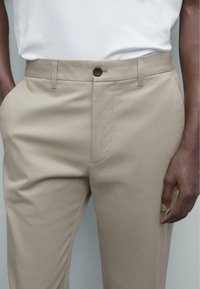 Beige tailored trousers made of smooth fabric, featuring a front zipper, button closure, and side pockets. Minimalist design, straight fit.