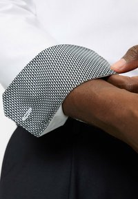 Weißes Hemd mit schwarzen gepunkteten Manschetten, die ein hexagonales Muster aufweisen. Die Manschette zeigt eine feine Textur und ein weißes Akzentdetail.