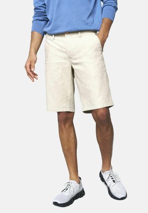 Mann trägt hellbeige knielange Shorts, ein blaues Langarmhemd und weiße Sneakers mit schwarzen Sohlen, steht mit einer Hand in der Tasche.