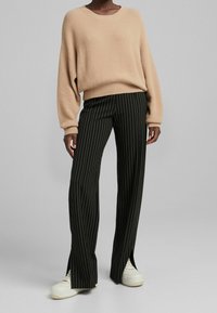 Personne portant un pull beige à manches longues, un pantalon large à fines rayures noires avec des fentes à l'avant, et des baskets blanches sur un fond gris uni.