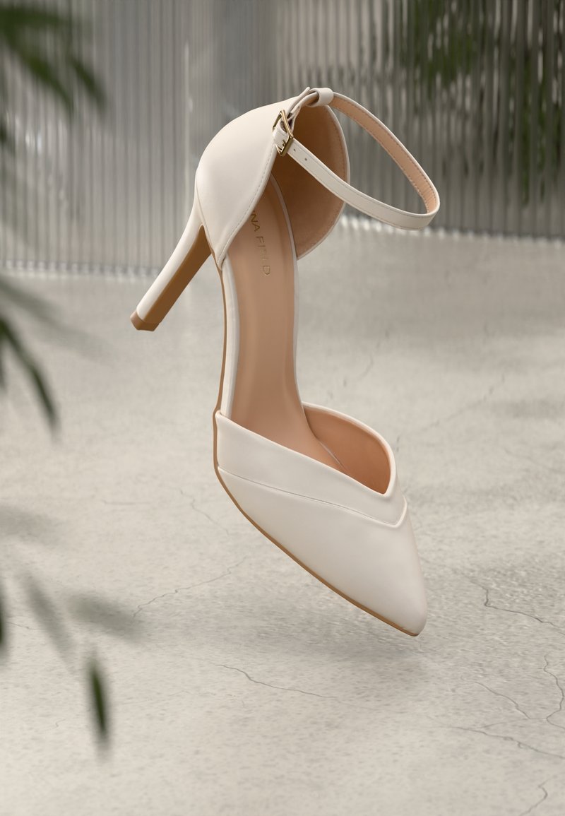 Chaussure à talon haut beige avec bout pointu et bride à la cheville, flottant au-dessus d'un sol en béton fissuré avec des plantes floues et un fond ondulé.