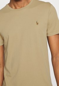 Vaaleanruskea lyhythiha t-paita, joka on valmistettu pehmeästä puuvillasta. Siinä on pieni brodeerattu logo vasemmassa rintakohdassa. Pyöreä pääntie.