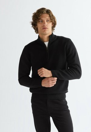 Jeune homme aux cheveux bouclés portant un pull noir à fermeture éclair mi-longue et un pantalon noir, ajustant sa manche gauche sur un fond clair uni.