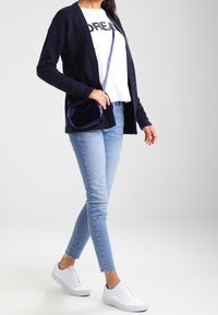 Marineblauer Cardigan, weißes T-Shirt mit schwarzem Text, hellblaue Skinny-Jeans und weiße Sneaker. Kleine marineblaue Umhängetasche setzt einen Akzent.