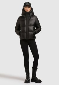 Kvinde i sort pufferjakke med hætte, sorte leggins, kraftige sorte støvler og sort baseball-cap med hvidt logo, der står mod grå baggrund.