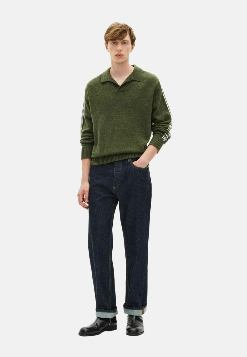 Un jeune homme se tient avec une main dans la poche, portant un pull en maille vert avec des détails rayés sur les manches, un jean foncé retroussé et des bottes noires.