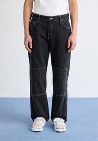 Pantaloni din denim negri cu cusături albe contrastante, design cu picioare largi și buzunare frontale. Purtați cu adidași bej pe un podea de un albastru deschis.