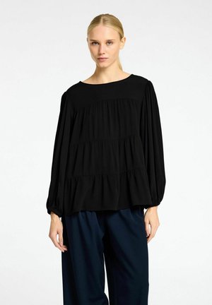 Sort bluse med lange ærmer, rund halsudskæring og lagdelt rufflesdesign. Blødt stof med samlede manchetter. Båret med mørkeblå bukser.