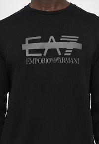 Svart langermet skjorte med et grått trykt logo "EA7 Emporio Armani" over brystet, med glatt tekstur og rund hals.