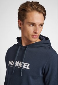 Marinblå huvtröja med dragsko, har en stor vit "HUMMEL"-logotyp och text. Mjuk bomullsmaterial, ergonomisk passform och inga synliga mönster.