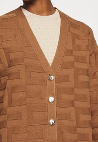 Cardigan marrón texturizado con escote en V, cuatro botones metálicos y un patrón de tejido. Llevado sobre una blusa de punto color crema.