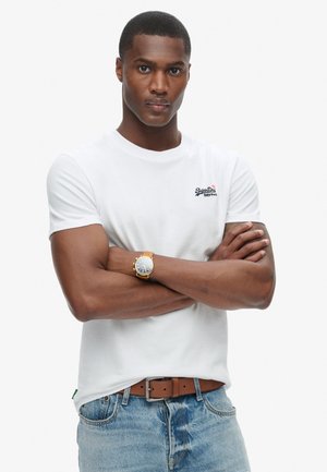 White crew-neck t-shirt with short sleeves, featuring a small logo on the chest. Paired with blue denim jeans and a brown belt.