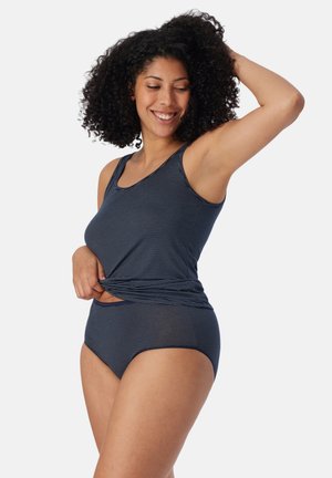 Femme aux cheveux bouclés portant un débardeur rayé bleu marine et une culotte taille haute assortie, souriant avec un bras levé.