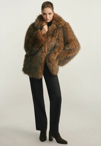 Brązowy sztuczny futrzany płaszcz z szerokim kołnierzem i grubą teksturą, zestawiony z czarnymi eleganckimi spodniami i botkami na kwadratowym obcasie.