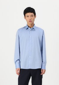 Cămașă albastră deschis cu mânecă lungă, dintr-un material neted, cu guler clasic, butoniere pe față și manșete. Asortată cu pantaloni în dungi întunecate.