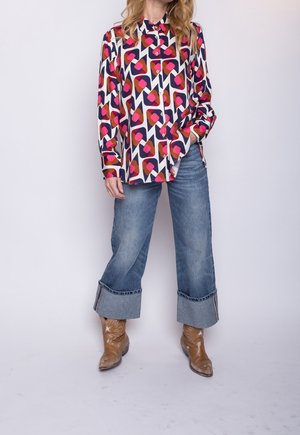 Vrouw draagt een kleurrijk overhemd met een geometrisch patroon, brede omgeslagen blauwe jeans en bruine westernlaarzen, staand tegen een effen achtergrond.