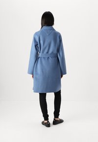 Abrigo largo de lana azul con cinturón, de corte oversize y gran cuello. La parte trasera presenta un detalle de parche triangular. Se lleva sobre leggings negros y zapatos.