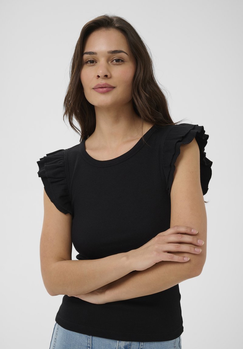 Jeune femme aux longs cheveux noirs portant un haut noir sans manches à volants et un jean bleu clair, les bras croisés, fond neutre.