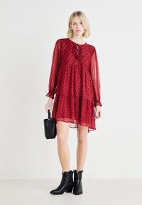Rochie roșie în straturi, cu decolteu rotund, mâneci lungi din materiale transparent, detalii cu șnur și mici modele albe. Purtați cu botine negre și o geantă.