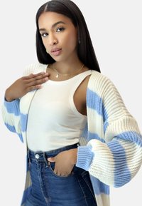 Stickad cardigan med blåa och vita ränder, mjuk textur, öppen front, ihopparad med en vit ribbad tanktop, högmidjade blå jeans.