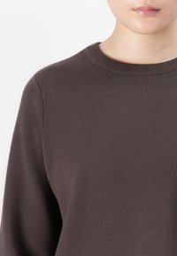 Person iført en mørk brun rundhals strikkegenser, som viser den øvre torso og en del av den nedre delen av ansiktet med lukte lepper.