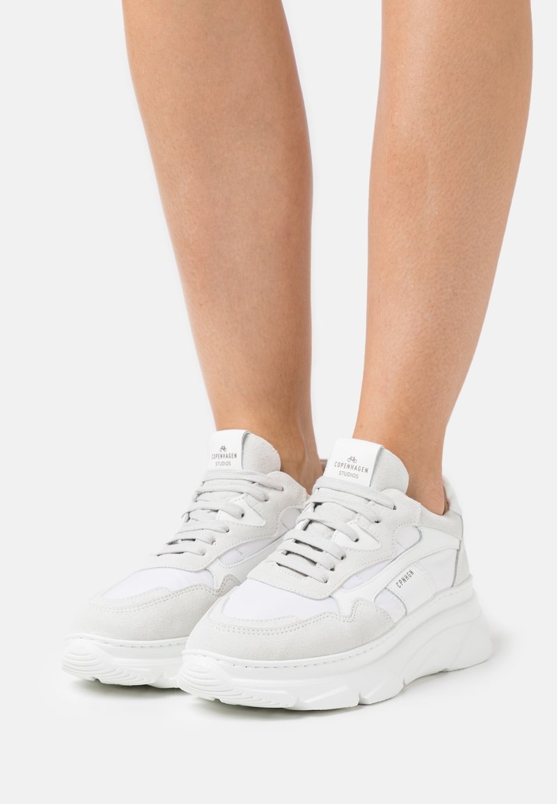 Füße in weißen und hellgrauen, klobigen Sneakers von Copenhagen Studios mit Schnürsenkeln auf weißer Oberfläche.