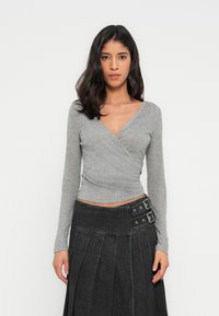 Γκρι μακρυμάνικη μπλούζα τύπου wrap με λαιμόκοψη σε σχήμα V, συνδυασμένη με μαύρη φούστα από τζιν με αγκράφες και πλισέ υφή.