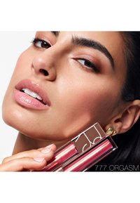 Læbefugtgivende gloss emballage har en klar, rektangulær beholder med en pink metallisk finish. Produktnavnet "777 ORGASM" er trykt på siden.
