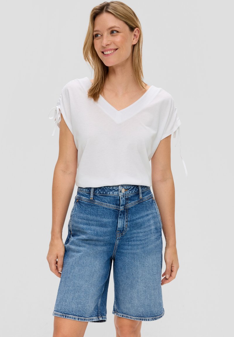 T-shirt bianca a collo a V con maniche corte arrotolate; abbinata a pantaloni Bermuda in denim blu, con leggeri segni di usura e orlo sfilacciato.