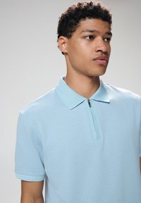 Polo shirt di colore azzurro chiaro con tessuto testurizzato, maniche corte e colletto con zip. Presenta un motivo a quadretti su tutta la superficie.