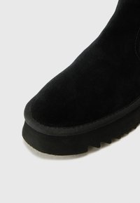 Bottine en suède noir avec un bout rond, une surface texturée et une semelle en caoutchouc crantée. Présente une bordure cousue sur le dessus.