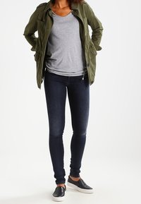 Chaqueta verde oliva, camiseta gris, jeans oscuros ajustados y zapatillas negras sin cordones. La chaqueta tiene bolsillos y cierre de botón.