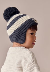 Gestrickte Mütze mit Streifen und Bommel, in Marineblau und Cremefarben, mit gerippter Struktur und Ohrenschützern. Verziert mit einem runden Aufnäher.
