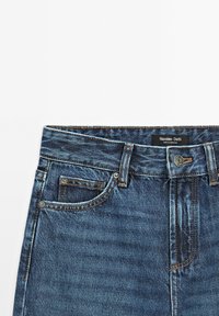 Jeans in denim blu scuro con zip frontale, design a cinque tasche e una patch in pelle sulla cintura posteriore. Sfumature e segni delicati.