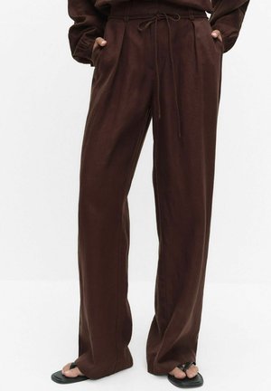 Pantalon marron ample avec taille à cordon, porté avec des sandales noires type tongs, mains dans les poches latérales.