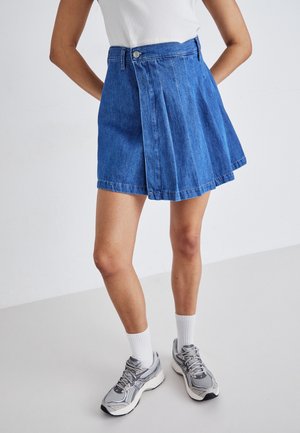 Kvinde iført blå denimnederdel med folder foran, hvid ribbet ærmeløs top, hvide ankelsokker og grå atletiksneakers.
