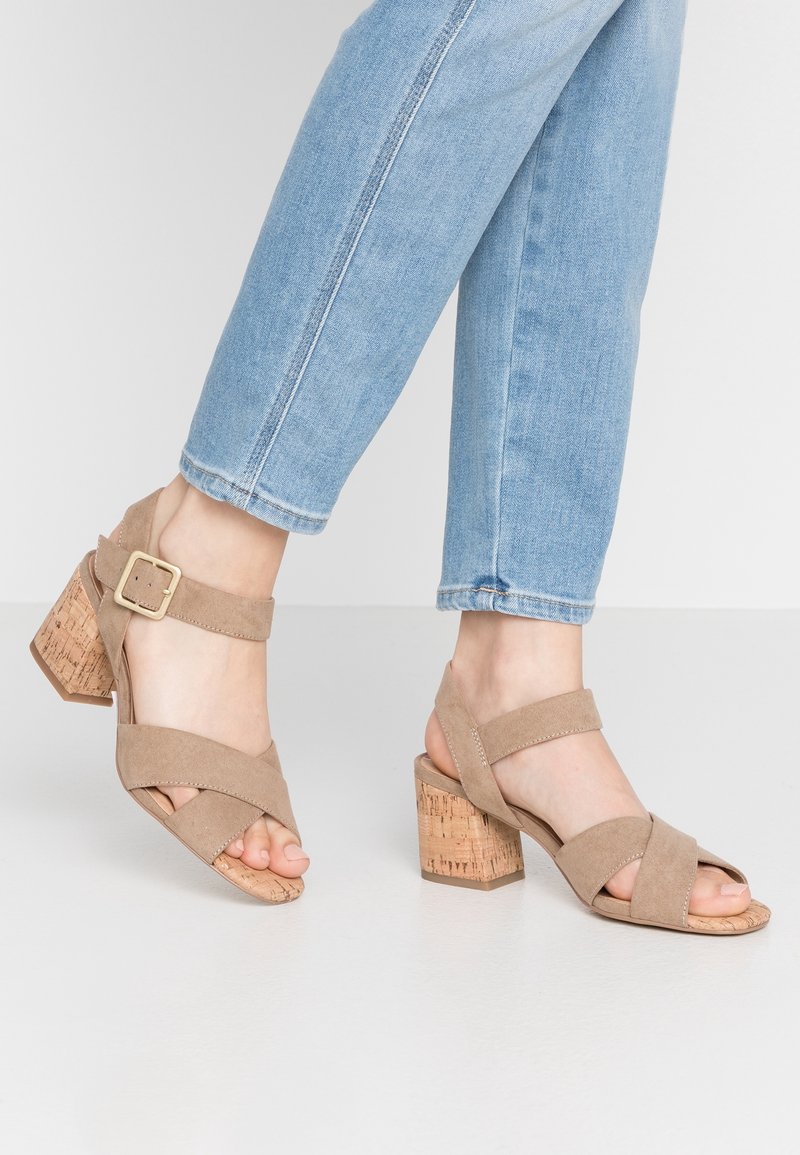 Pieds portant des sandales à talons blocs en daim beige avec des brides autour de la cheville et des boucles dorées, associées à un jean en denim bleu clair.