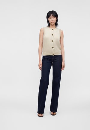 Kvinde iført en ærmeløs beige knapvest med mørkeblå jeans med lige ben og sorte sandaler med åben tå, stående på en ensfarvet baggrund.