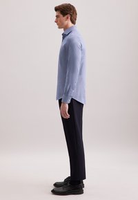 Man in een lichtblauw gestreept overhemd met lange mouwen, zwarte broek en zwarte nette schoenen, staande in profiel tegen een effen achtergrond.