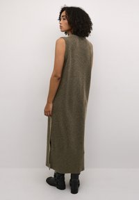 Robe longue sans manches en tissu tricot olive. Dotée d'entailles sur les côtés et d'un ourlet côtelé. Portée avec des bottines noires.