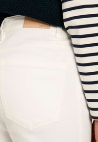 Valged teksad sileda tekstuuriga, tagaküljel on tasku ja beež nahktagune silt, millel on embossed tekst "JOULES". Riba äärisega pika varrukaga pluus on osaliselt nähtav.