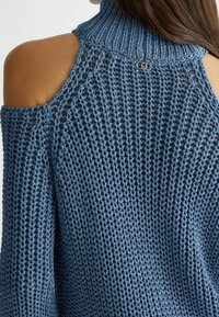 Pull bleu tricoté avec un col haut et un design à épaules dénudées. Présente un motif texturé et un accent en matériel argenté à l'arrière.