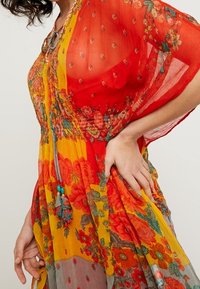 Abito floreale in rosso, giallo e grigio con tessuto trasparente. Presenta una vita arricciata, maniche a campana e dettagli decorativi con nappine.