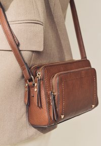 Sac bandoulière en cuir marron avec plusieurs compartiments zippés, accents métalliques dorés, et finition texturée, porté sur un fond en tissu beige.