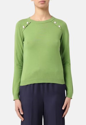 Persona che indossa un maglione verde a maniche lunghe con bottoni argentati sulle spalle e pantaloni blu scuro, in piedi davanti a uno sfondo semplice.